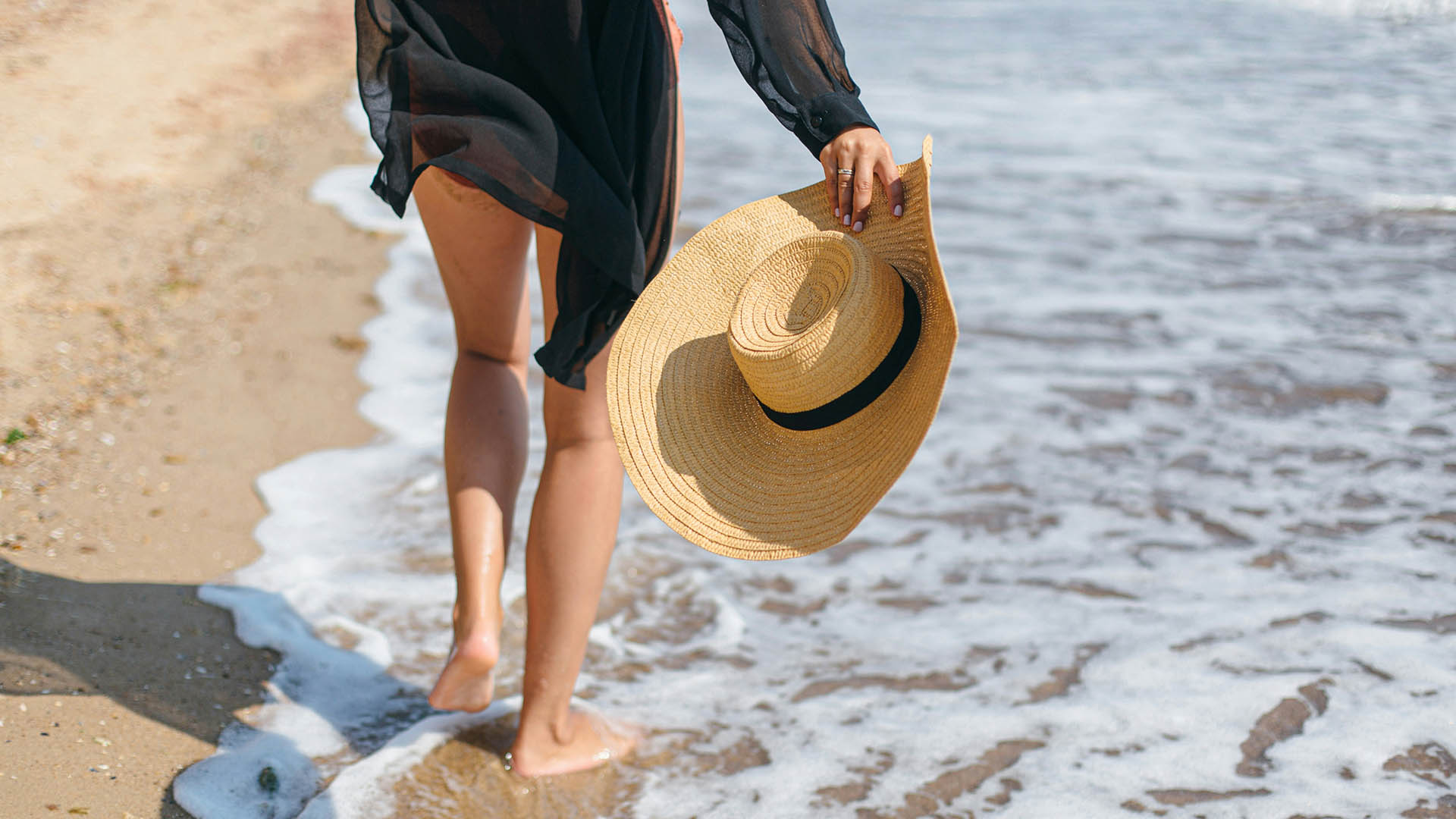 Person carrying straw sun hat in hand walking along sandy beach.
