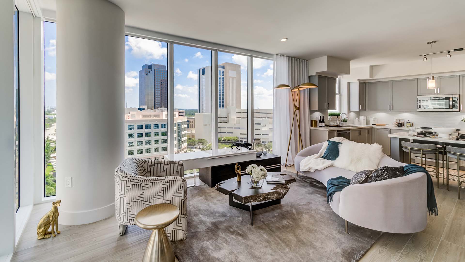 Apartment living area with large windows, plush modern furniture, and contemporary kitchen.